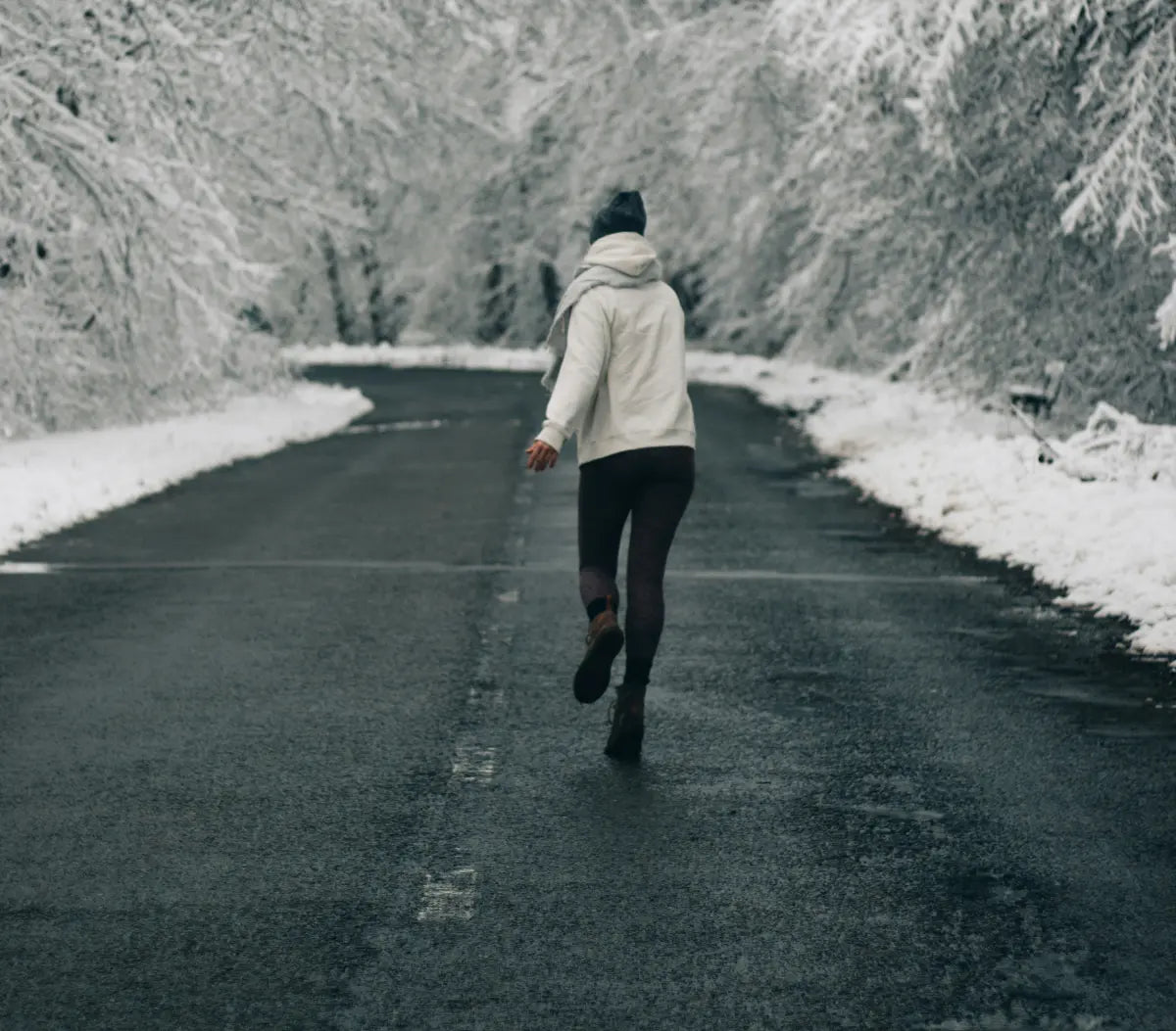 Henkilö talvivaatteissa juoksee tyhjää asvalttitietä kohti lumisia puita ja oksia molemmin puolin.