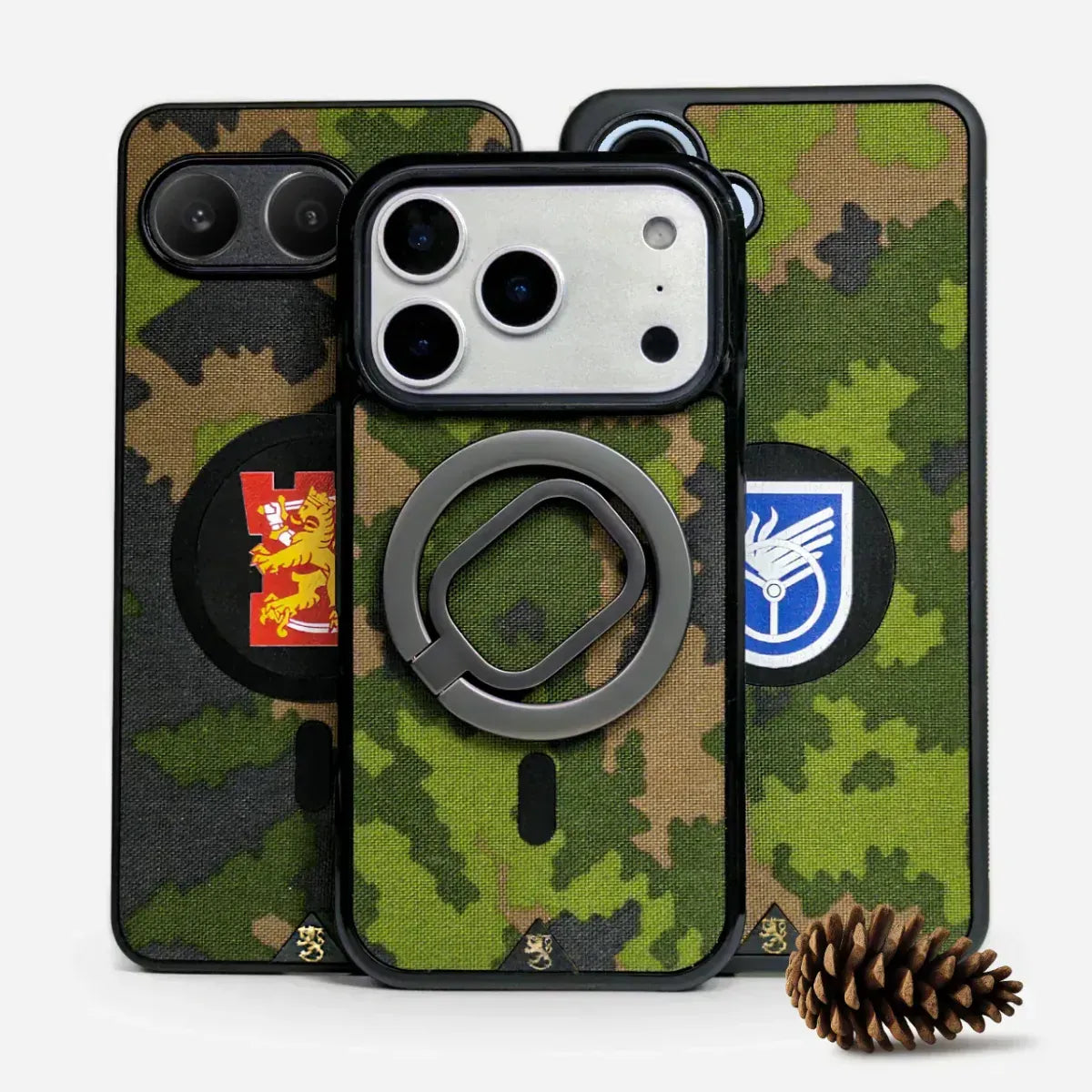 Kolme MagSafe-yhteensopivaa tekstiilipintaista armeijakuviollista puhelinkuorta, keskellä sormirenkaita ja vaakunakuvioita.