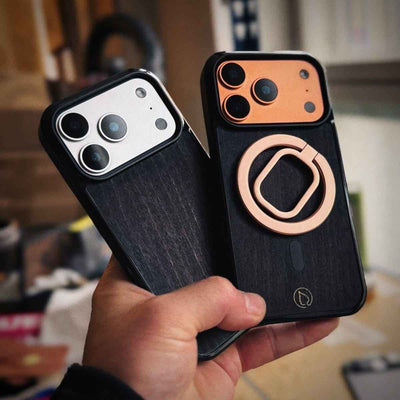Lastu  Käsi pitää kiinni kahdesta Lastu HIILI -merkkisestä mustasta koivupuusta valmistetusta suojakuoresta, jotka on tarkoitettu iPhone 17 Pro- ja Galaxy S26 -malleille, sekä metallisesta kamerarenkaasta.