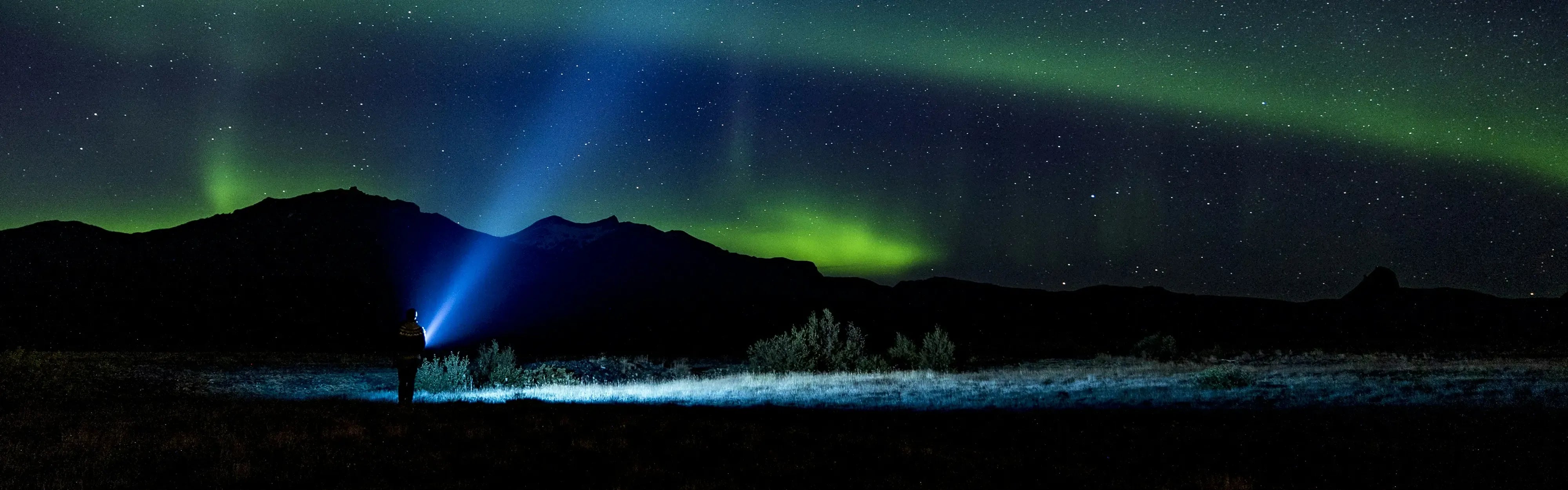 Yötaivaan revontulet kirkkaalla tähtitaivaalla, henkilö taskulampun valokeilassa tunturimaisemassa.